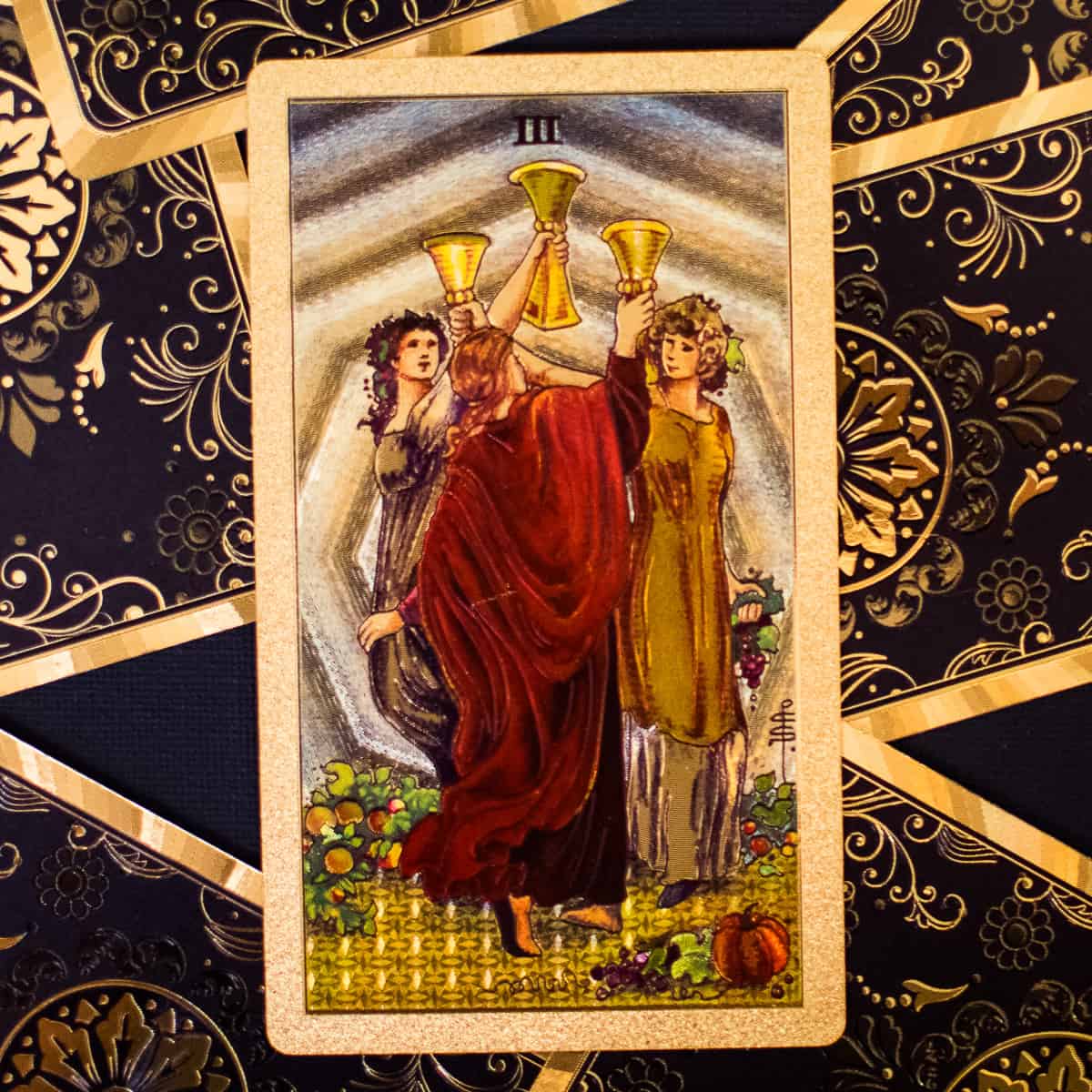 Three women celebrating with 3 gold cups depicted on a tarot card.
