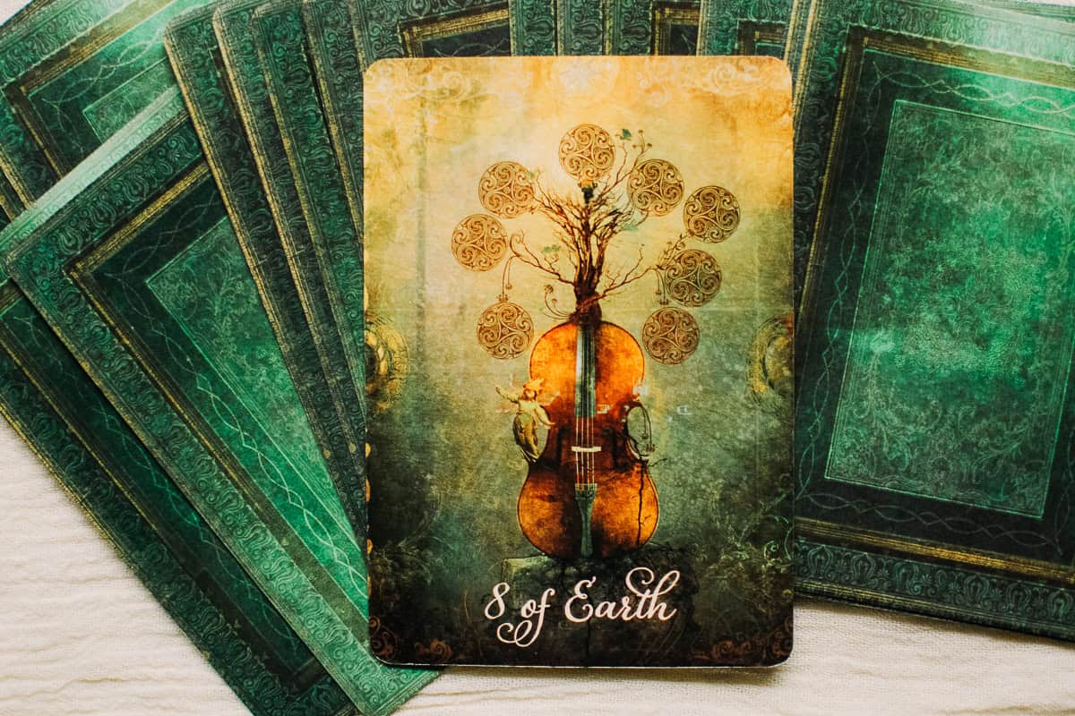 The Eight of Pentacles (Earth) card shown as a string instrument, perhaps a cello, surrounded by 8 pentacles. 