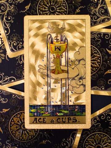 Hand holding overflowing cup of water on Ace of Cups tarot card.